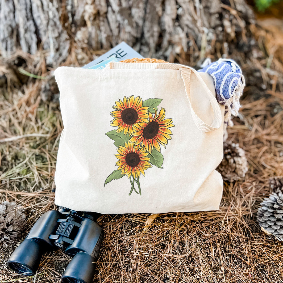 Canvas tote bag printed with a full-color illustration of a bunch of yellow and orange sunflowers, resting on dry grass beside a pair of binoculars.