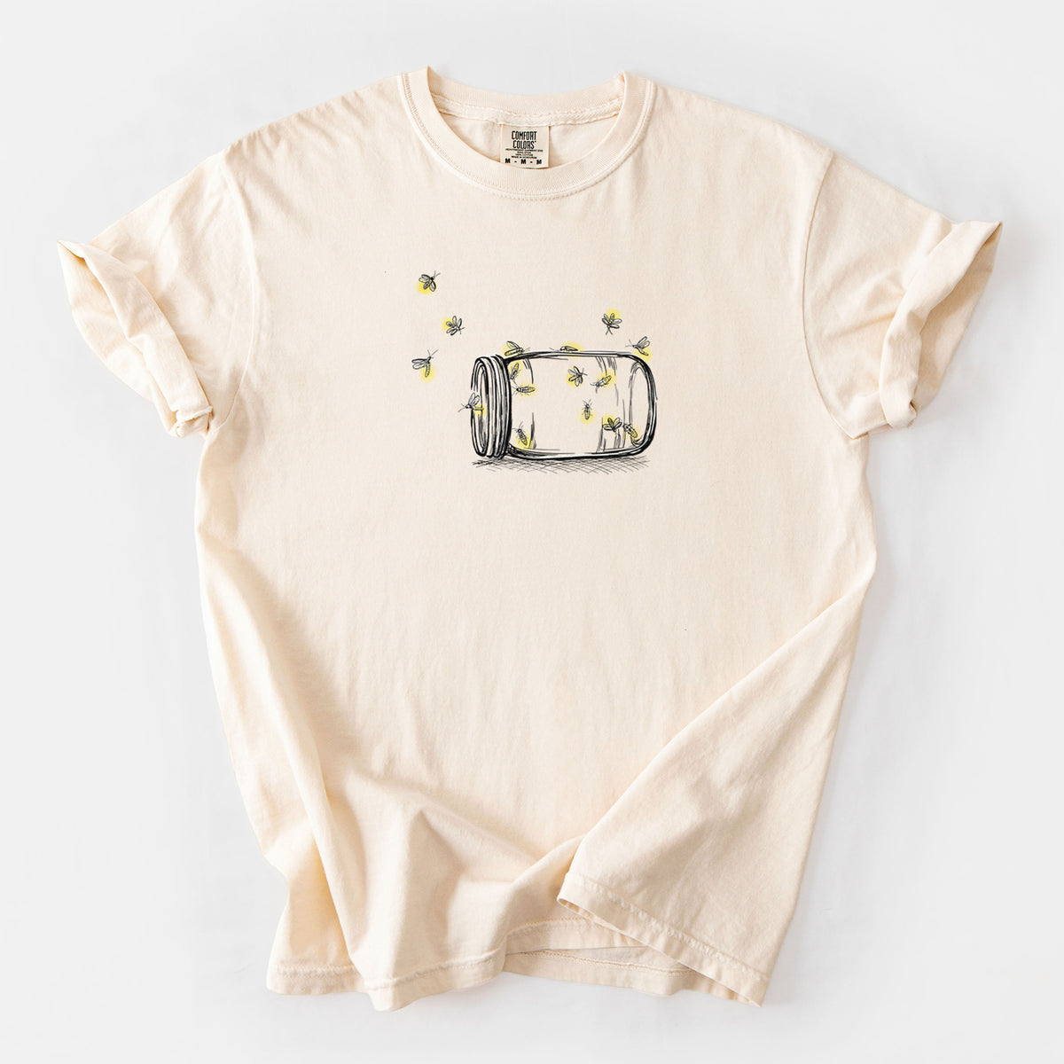 Ivory Comfort Colors t-shirt laid flat, printed with a mason jar with glowing yellow fireflies flying out of it.