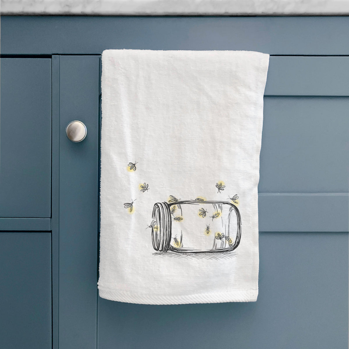 White hand towel printed with a mason jar with glowing yellow fireflies flying out of it, hanging on a cabinet door.