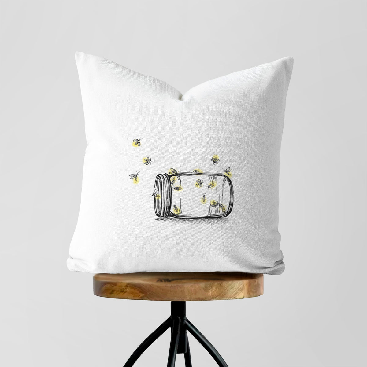 White pillow cover printed with a mason jar with glowing yellow fireflies flying out of it, on a wooden stool.