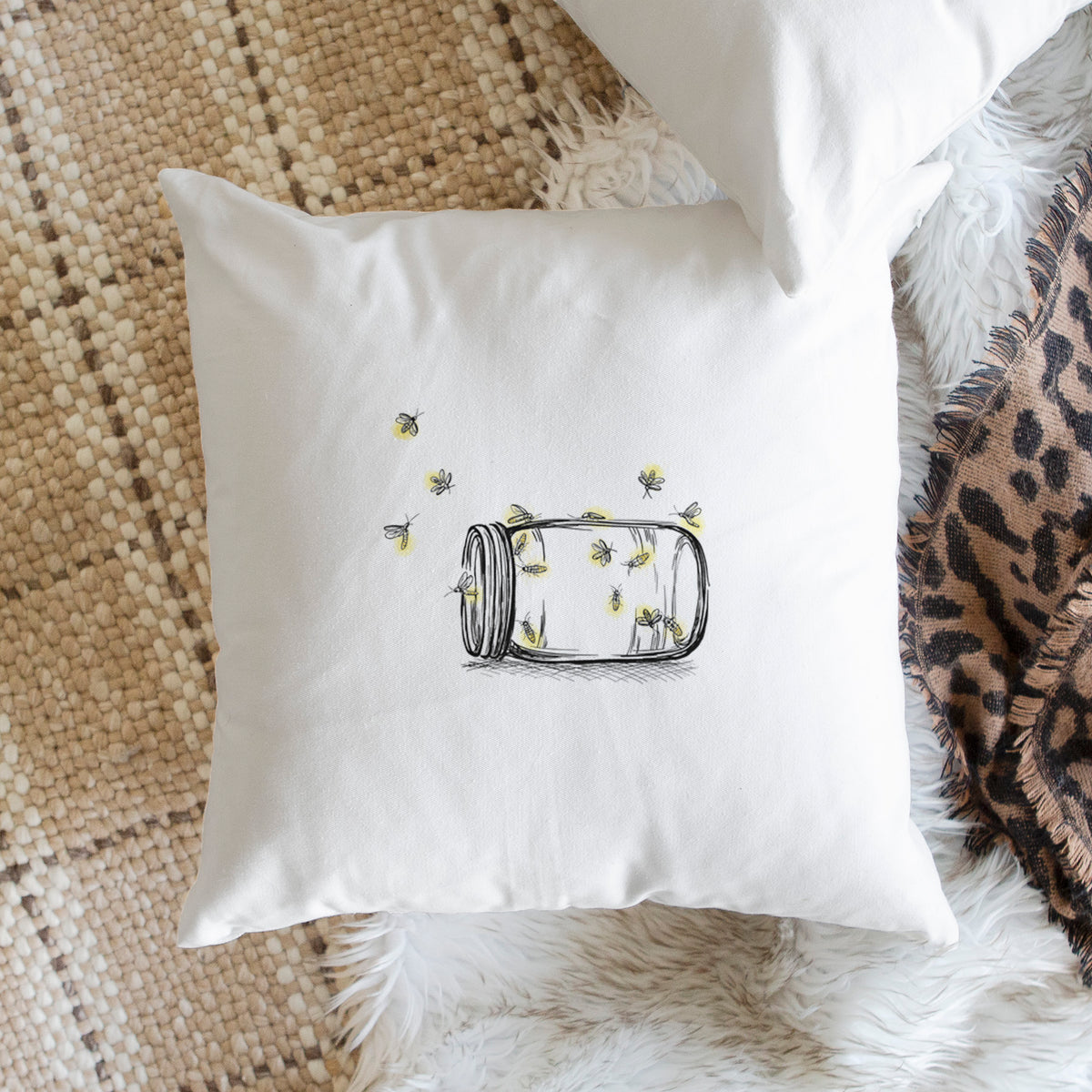 White pillow cover printed with a mason jar with glowing yellow fireflies flying out of it, on a woven chair.
