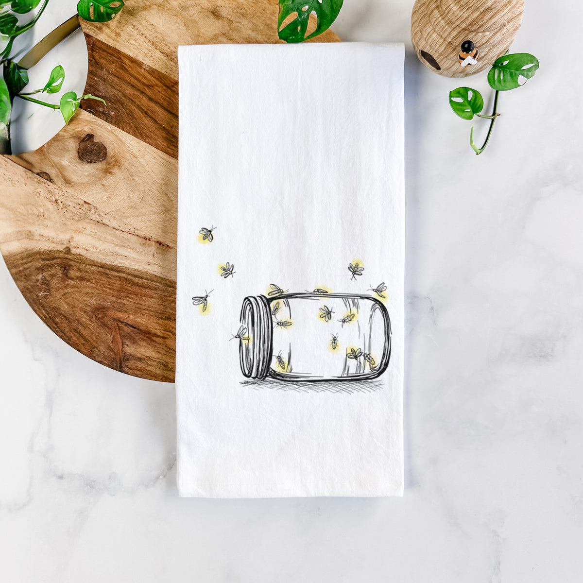 White cotton tea towel printed with a mason jar with glowing yellow fireflies flying out of it, on a marble counter beside a round wooden cutting board and greenery.