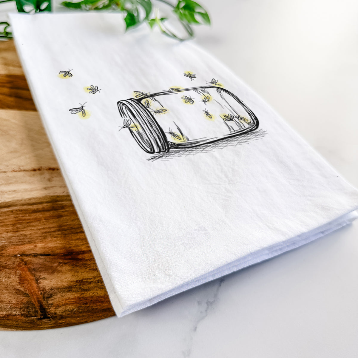 Close-up of a folded white tea towel on a wooden board, showing a mason jar with glowing yellow fireflies flying out of it.