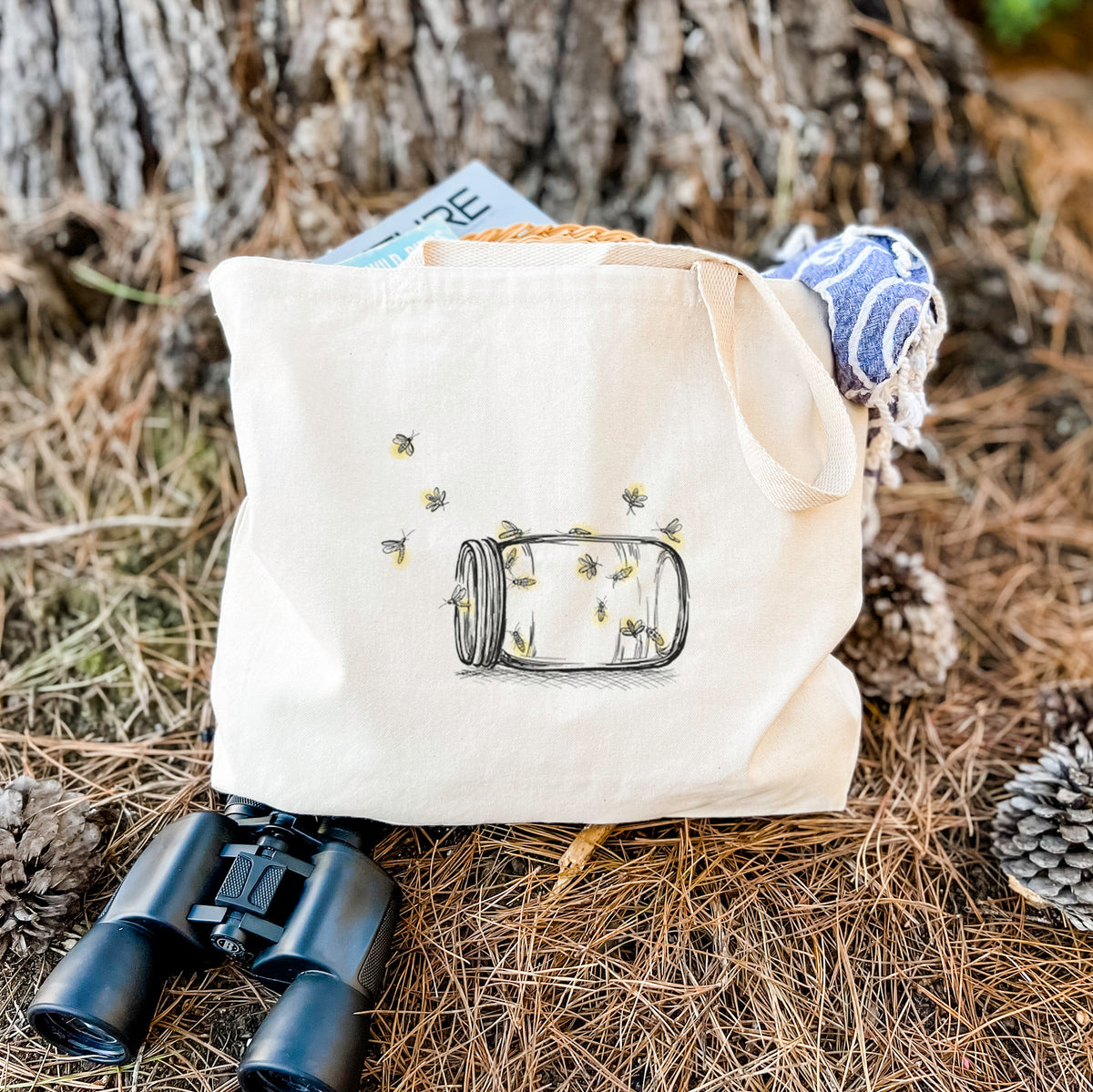 Canvas tote bag printed with a mason jar with glowing yellow fireflies flying out of it, resting on dry grass beside a pair of binoculars.