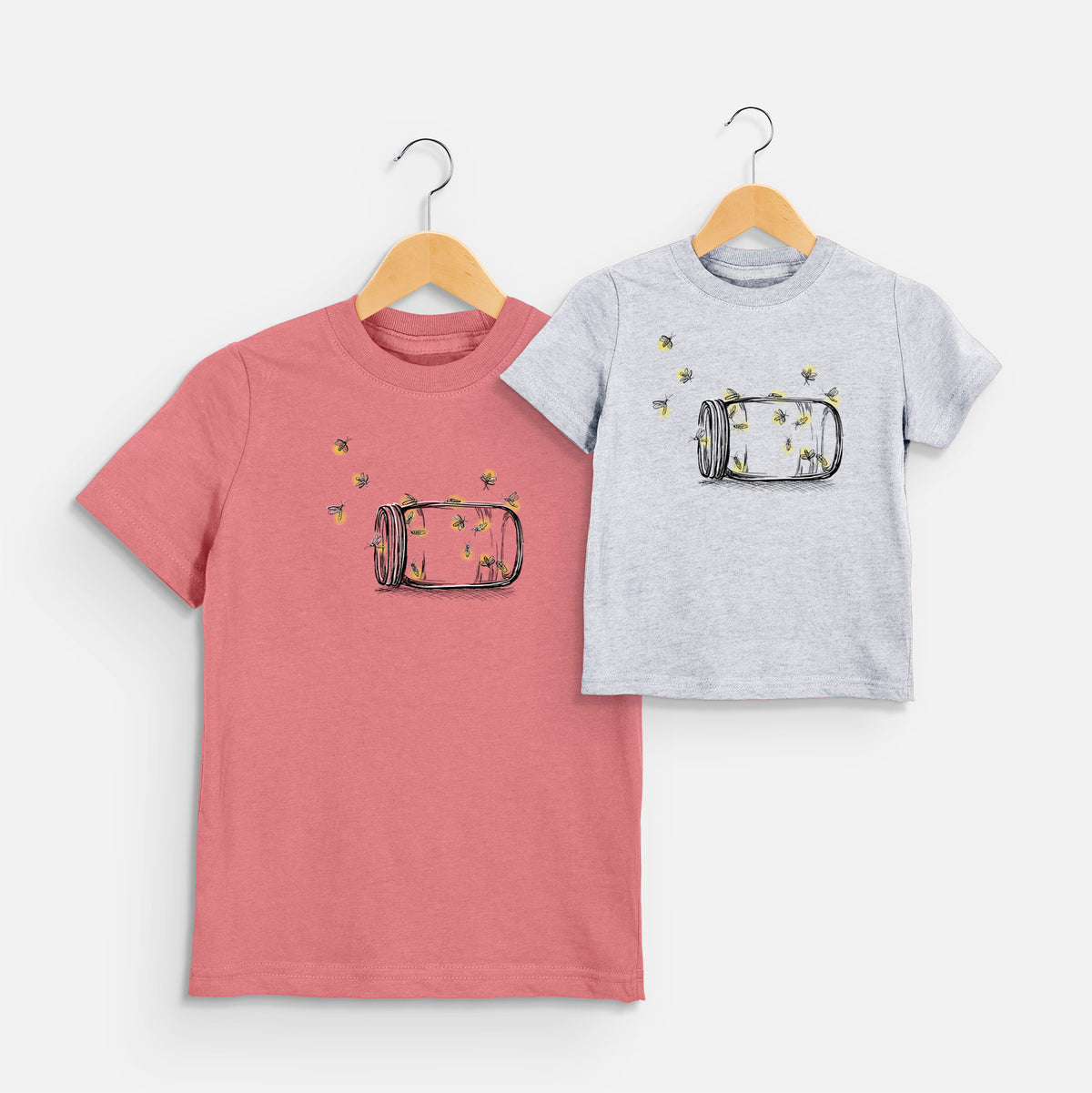Two youth t-shirts in different colors on hangers, printed with a mason jar with glowing yellow fireflies flying out of it.