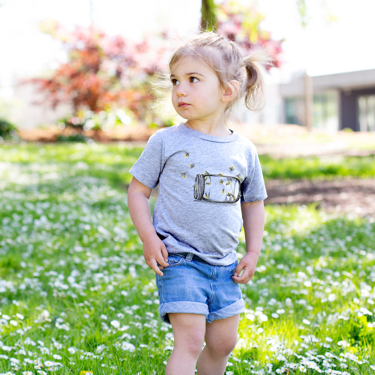 Toddler girl walking outdoors wearing a t-shirt printed with a mason jar with glowing yellow fireflies flying out of it.