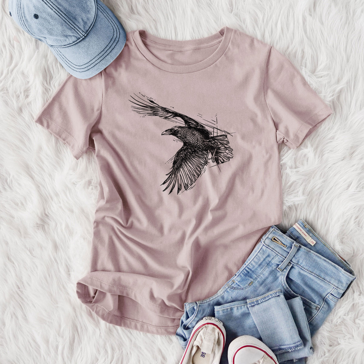 Women's relaxed fit cotton t-shirt printed with a black ink illustration of a raven in flight with its wings spread, styled flat on a white faux fur rug with jeans.