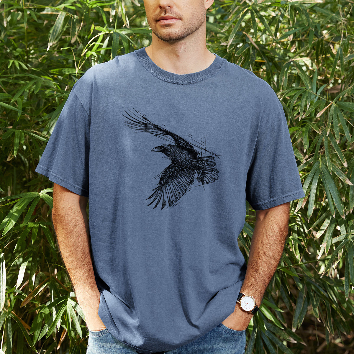 Man wearing a Comfort Colors t-shirt printed with a black ink illustration of a raven in flight with its wings spread, outdoors.