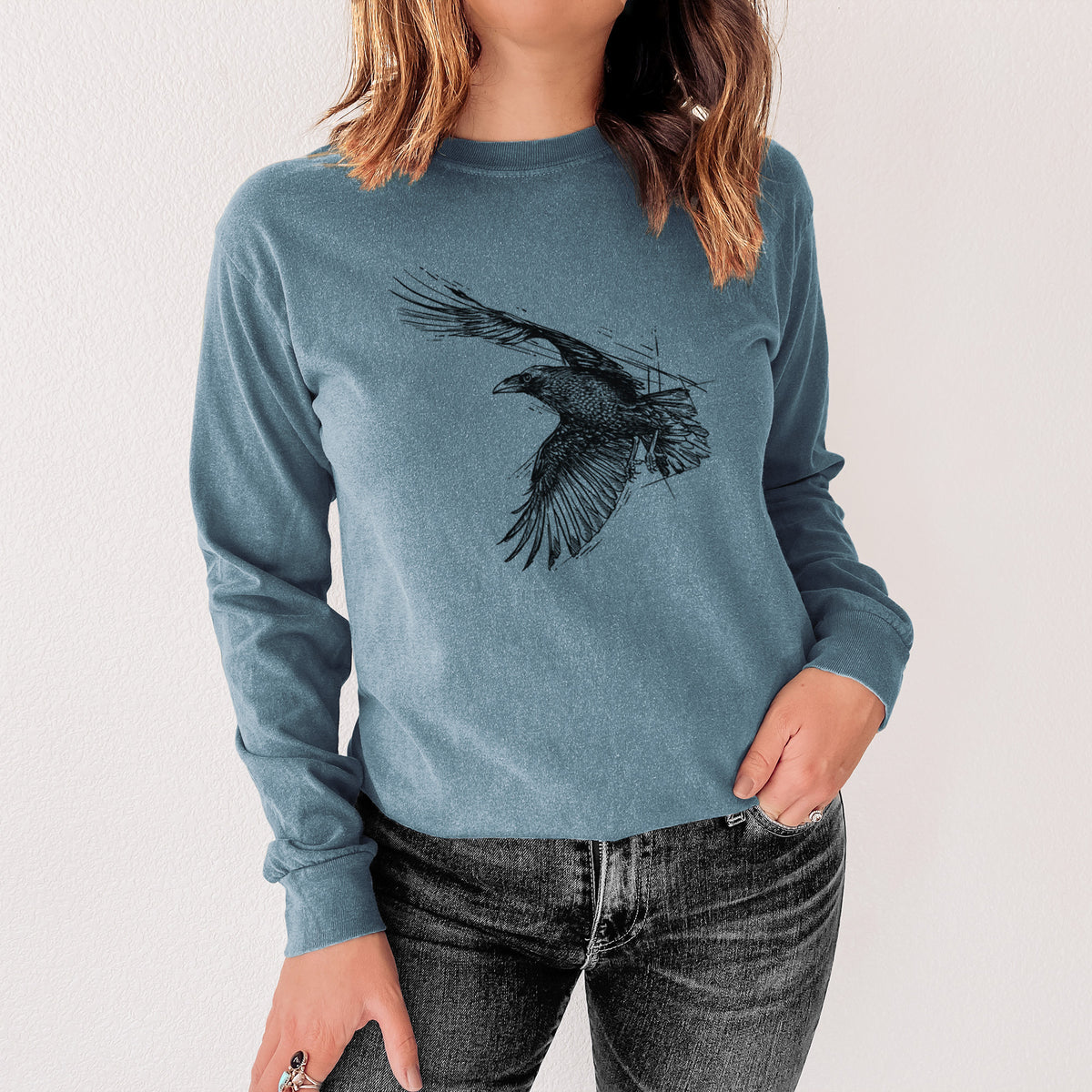 Steel blue Comfort Colors long sleeve shirt printed with a black ink illustration of a raven in flight with its wings spread, shown in a lifestyle photo.