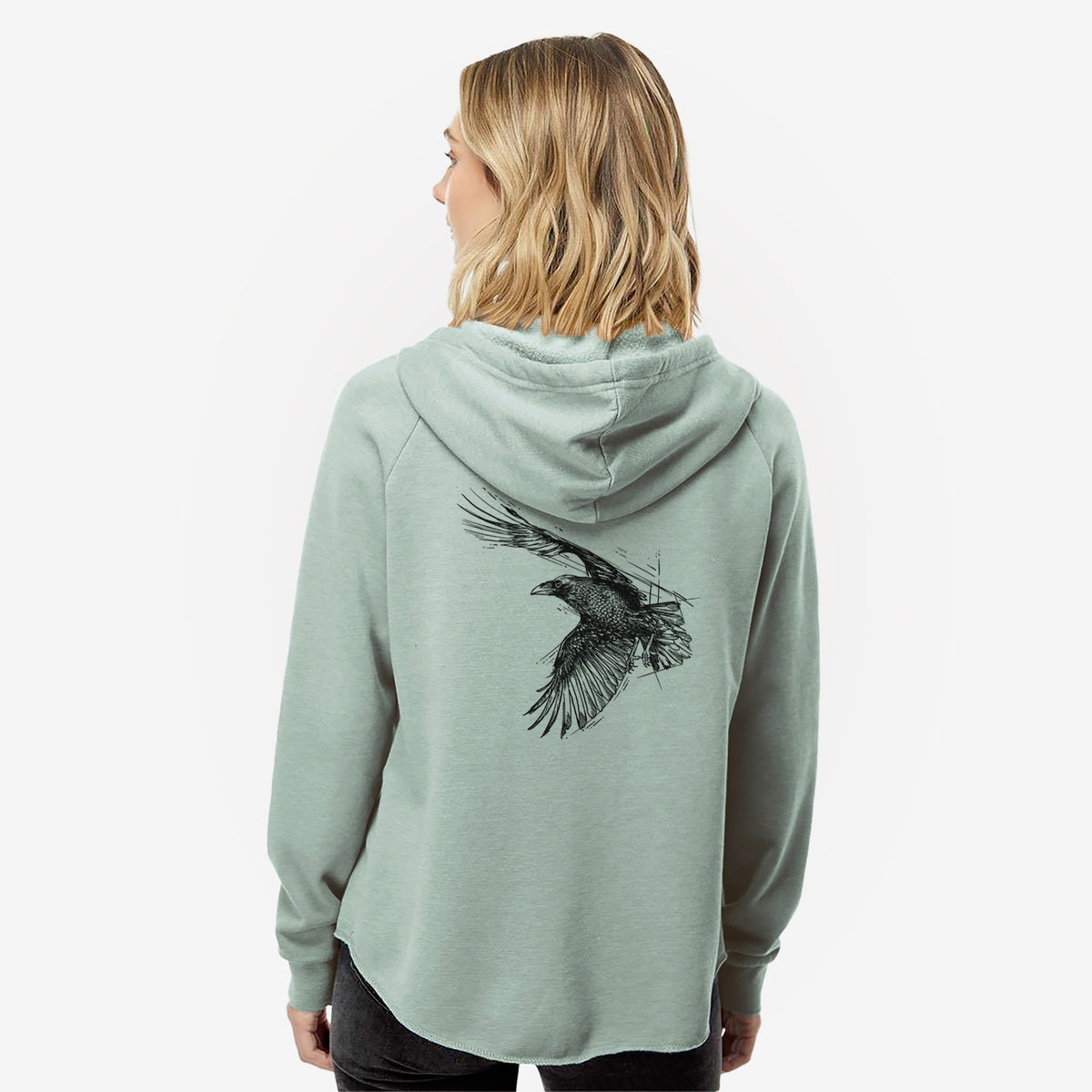 Woman wearing a sage green women's Cali Wave zip-up sweatshirt printed with a black ink illustration of a raven in flight with its wings spread, seen from behind.