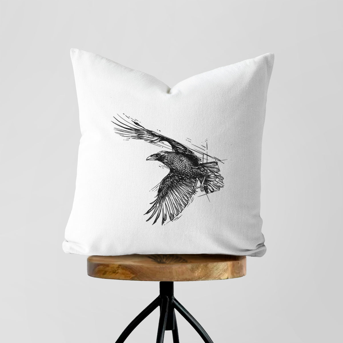 White pillow cover printed with a black ink illustration of a raven in flight with its wings spread, on a wooden stool.