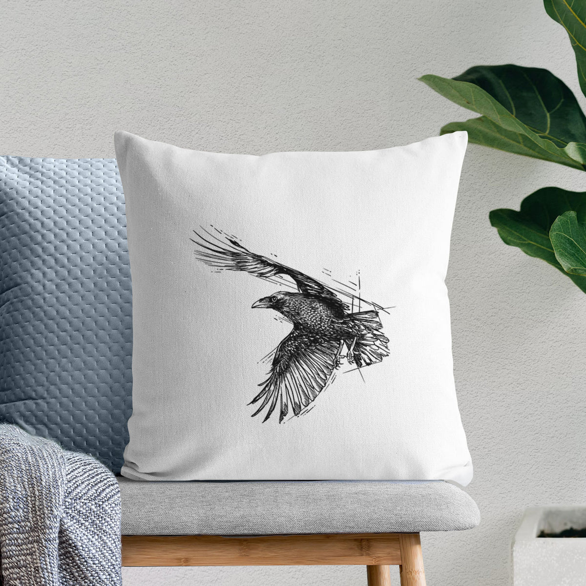 White pillow cover printed with a black ink illustration of a raven in flight with its wings spread, on a bench beside plants.