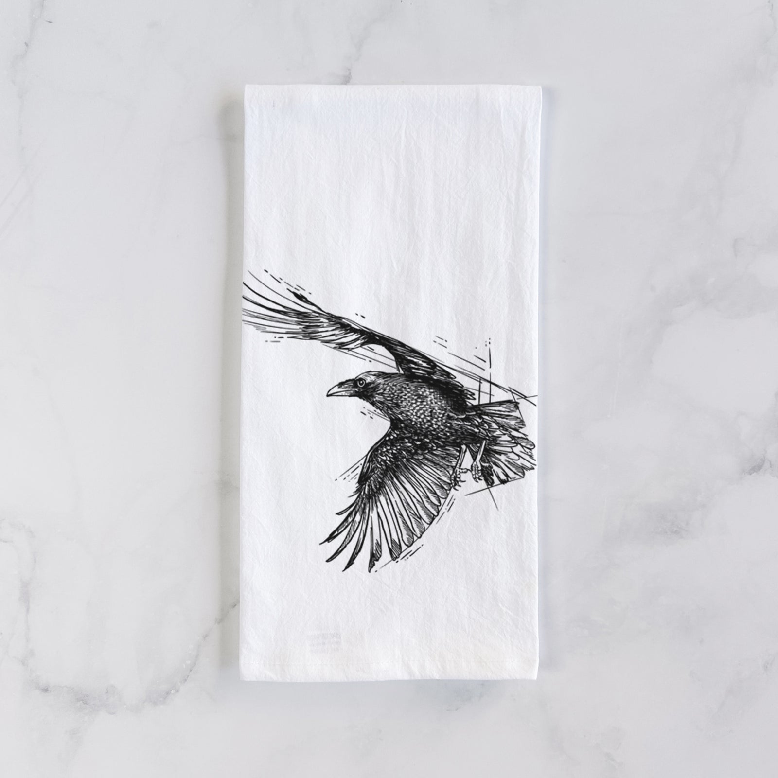 White cotton tea towel printed with a black ink illustration of a raven in flight with its wings spread, on a marble counter beside a round wooden cutting board and greenery.