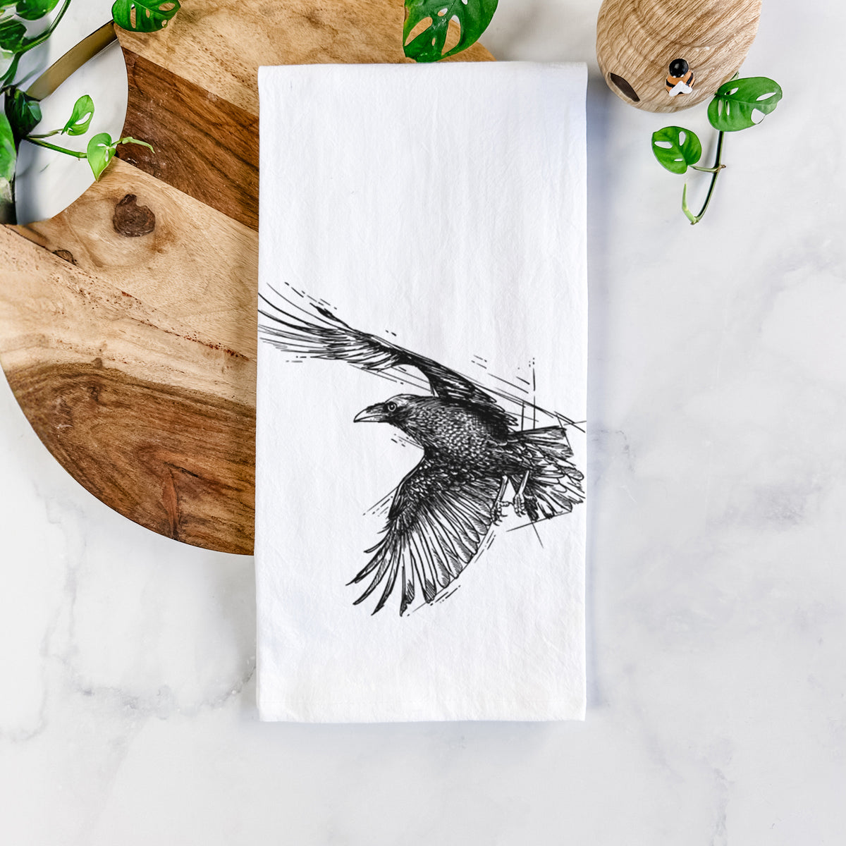 White cotton tea towel printed with a black ink illustration of a raven in flight with its wings spread, on a marble counter beside a round wooden cutting board and greenery.