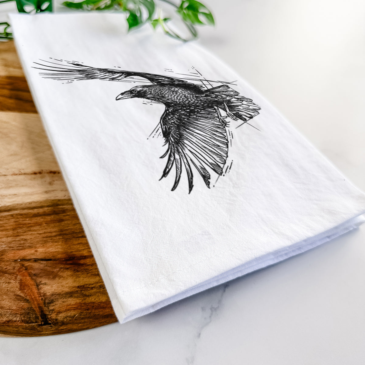Close-up of a folded white tea towel on a wooden board, showing a black ink illustration of a raven in flight with its wings spread.
