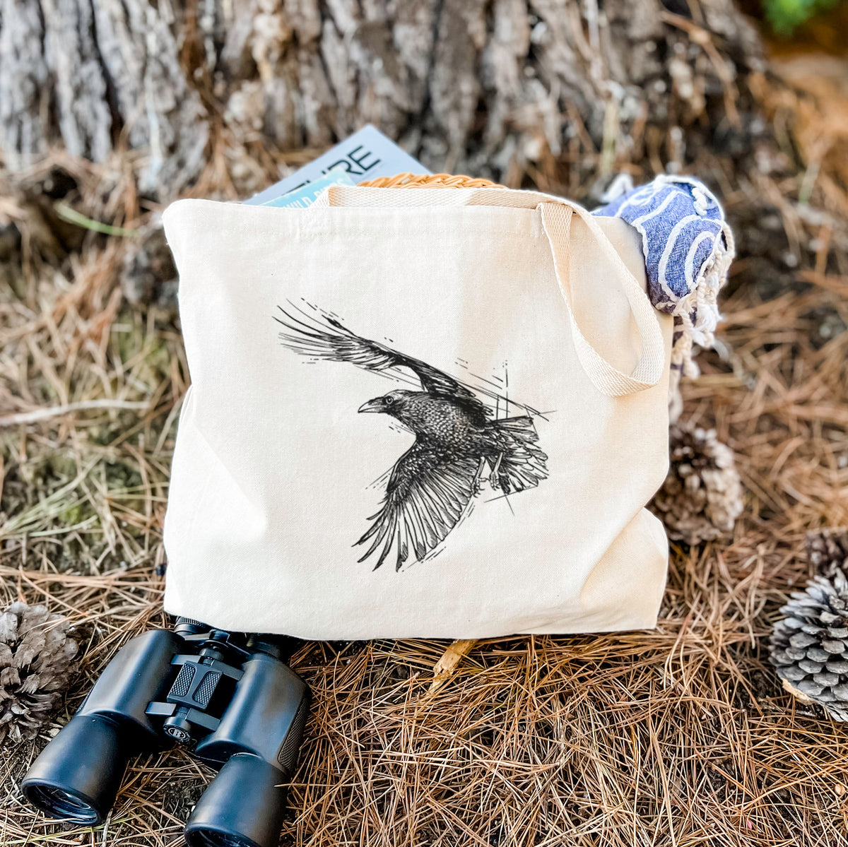 Canvas tote bag printed with a black ink illustration of a raven in flight with its wings spread, resting on dry grass beside a pair of binoculars.