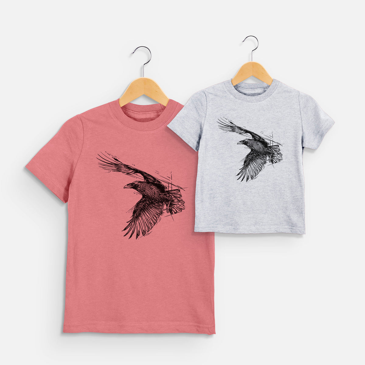 Two youth t-shirts in different colors on hangers, printed with a black ink illustration of a raven in flight with its wings spread.