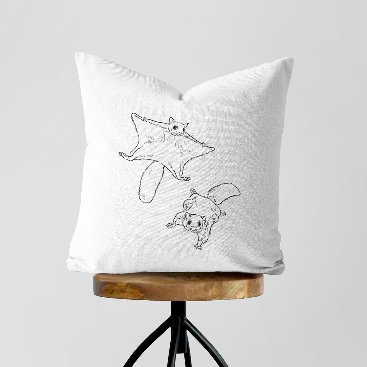 White pillow cover printed with a black ink illustration of two flying squirrels gliding with their skin flaps spread, on a wooden stool.