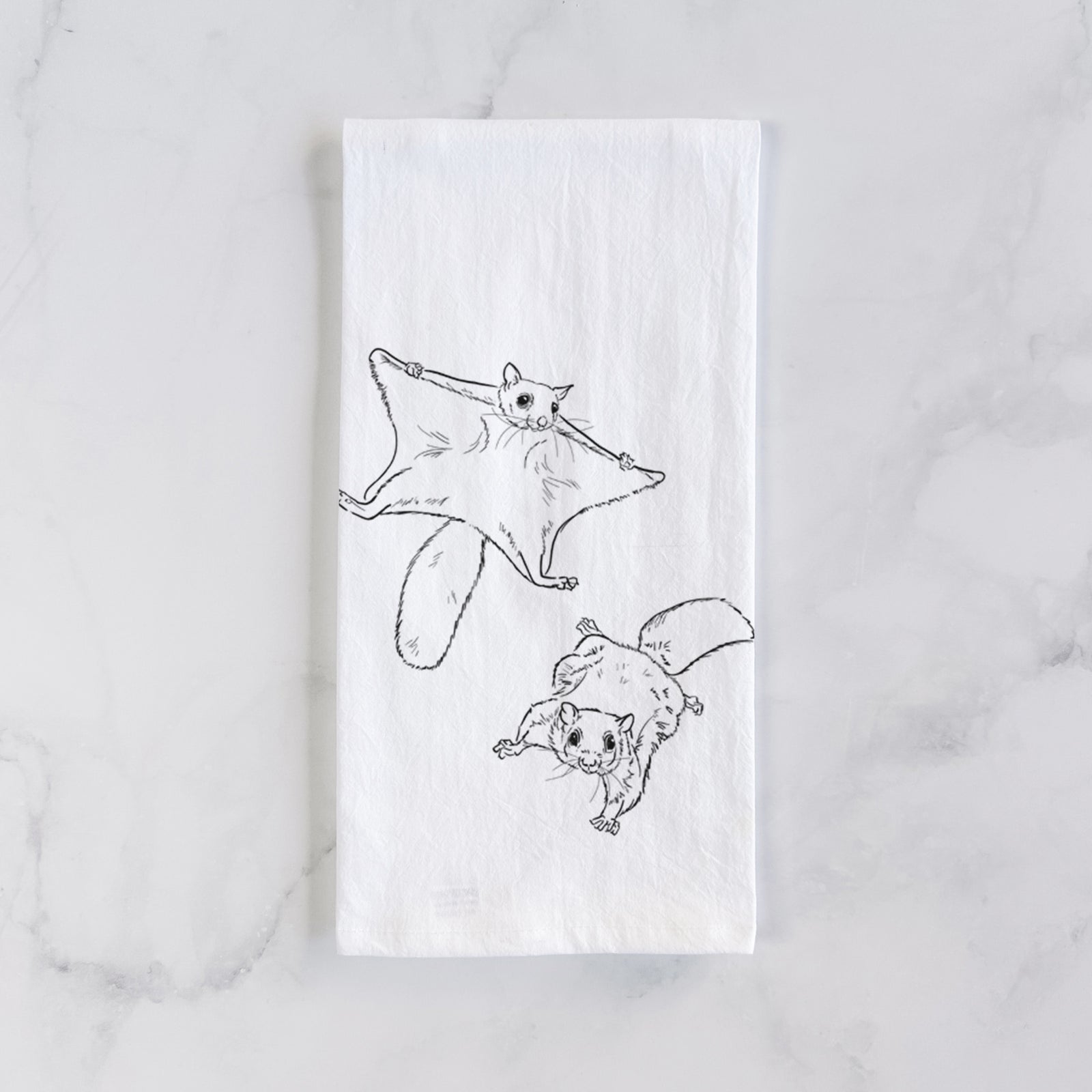 White cotton tea towel printed with a black ink illustration of two flying squirrels gliding with their skin flaps spread, on a marble counter beside a round wooden cutting board and greenery.
