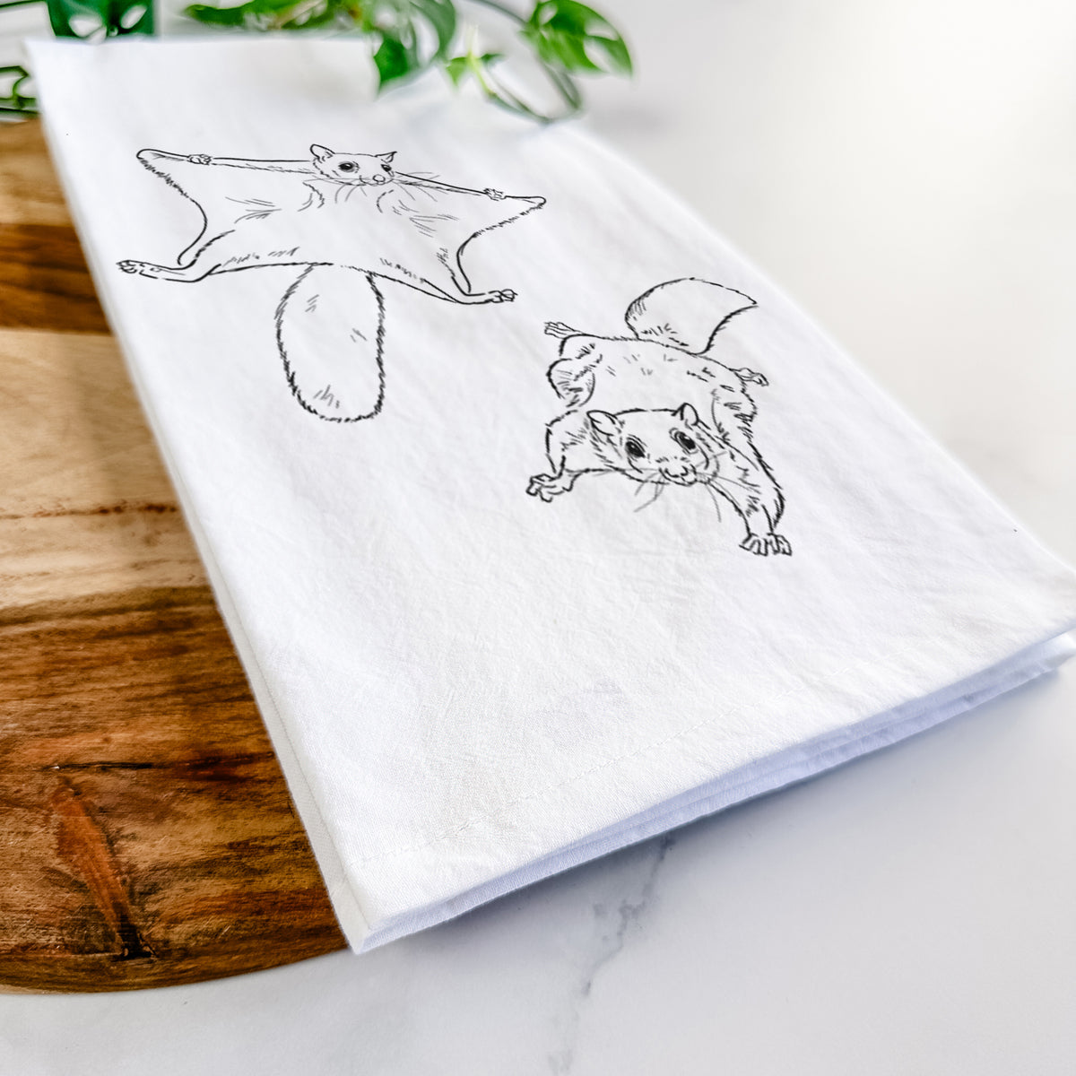 Close-up of a folded white tea towel on a wooden board, showing a black ink illustration of two flying squirrels gliding with their skin flaps spread.