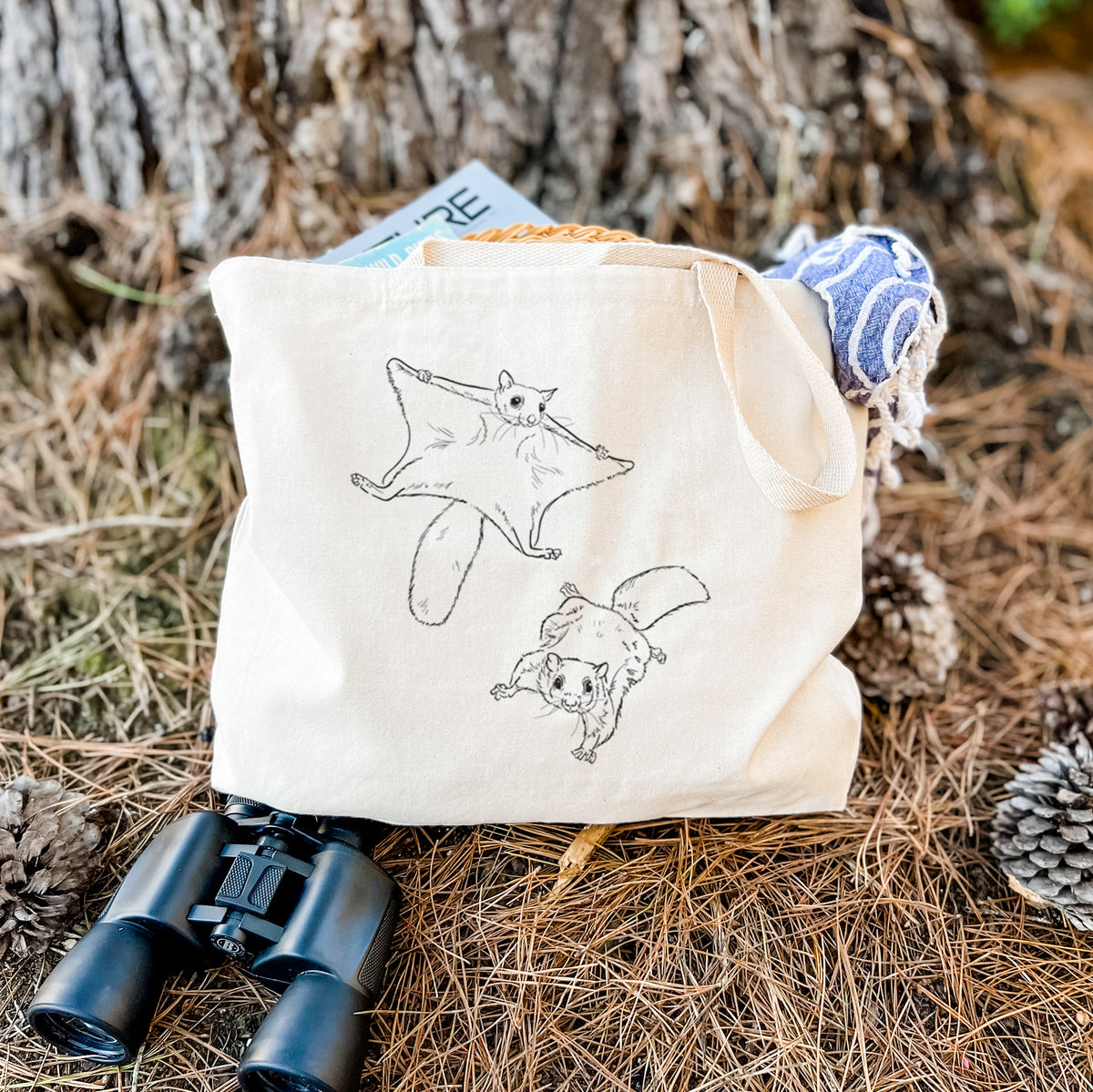 Canvas tote bag printed with a black ink illustration of two flying squirrels gliding with their skin flaps spread, resting on dry grass beside a pair of binoculars.
