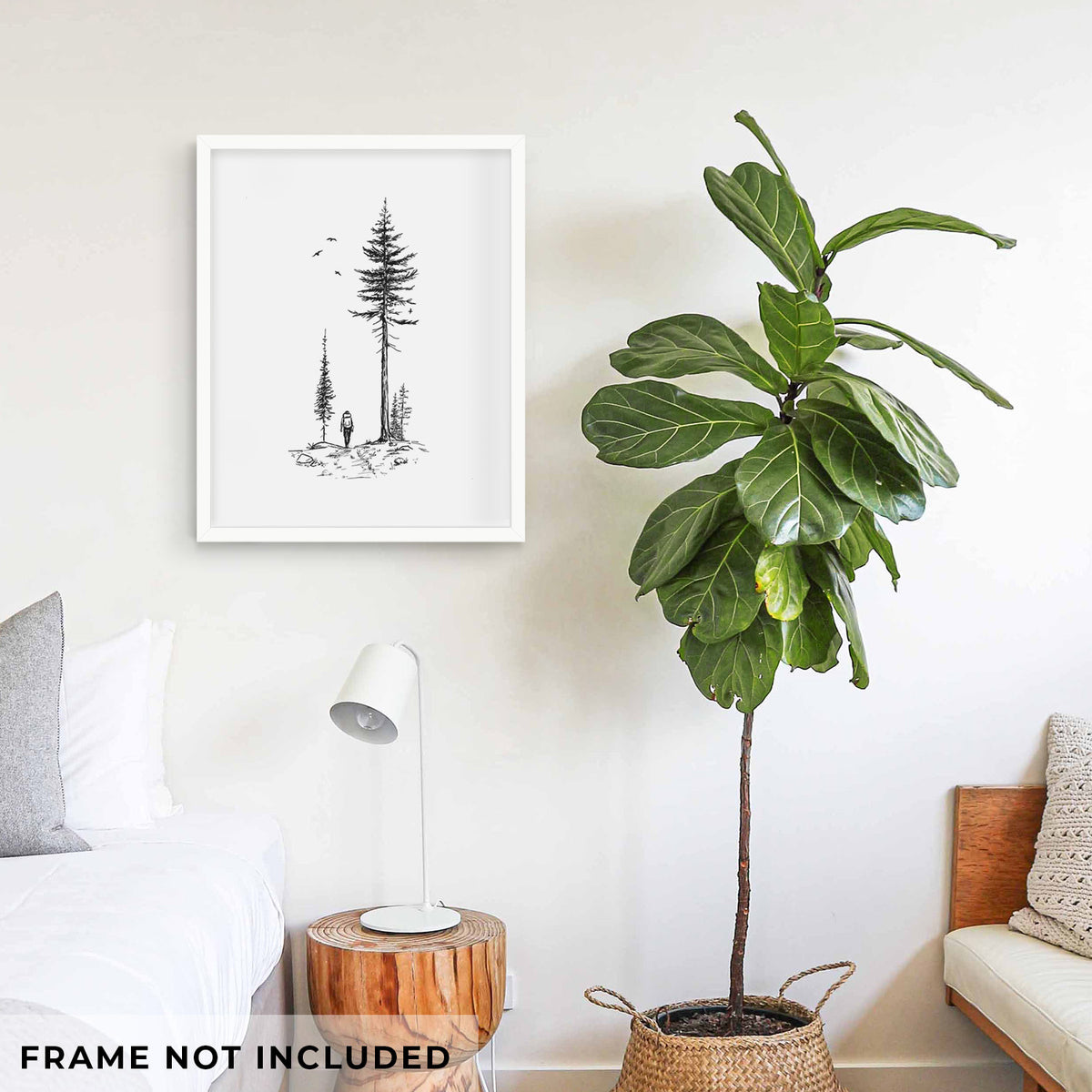 Art print of a black ink illustration of a lone hiker walking between tall pine trees with birds overhead, shown framed on a living room wall beside a potted plant. Frame not included.