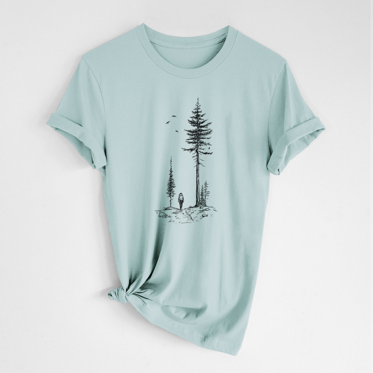Dusty blue lightweight cotton t-shirt laid flat with the sleeves rolled, printed with a black ink illustration of a lone hiker walking between tall pine trees with birds overhead.