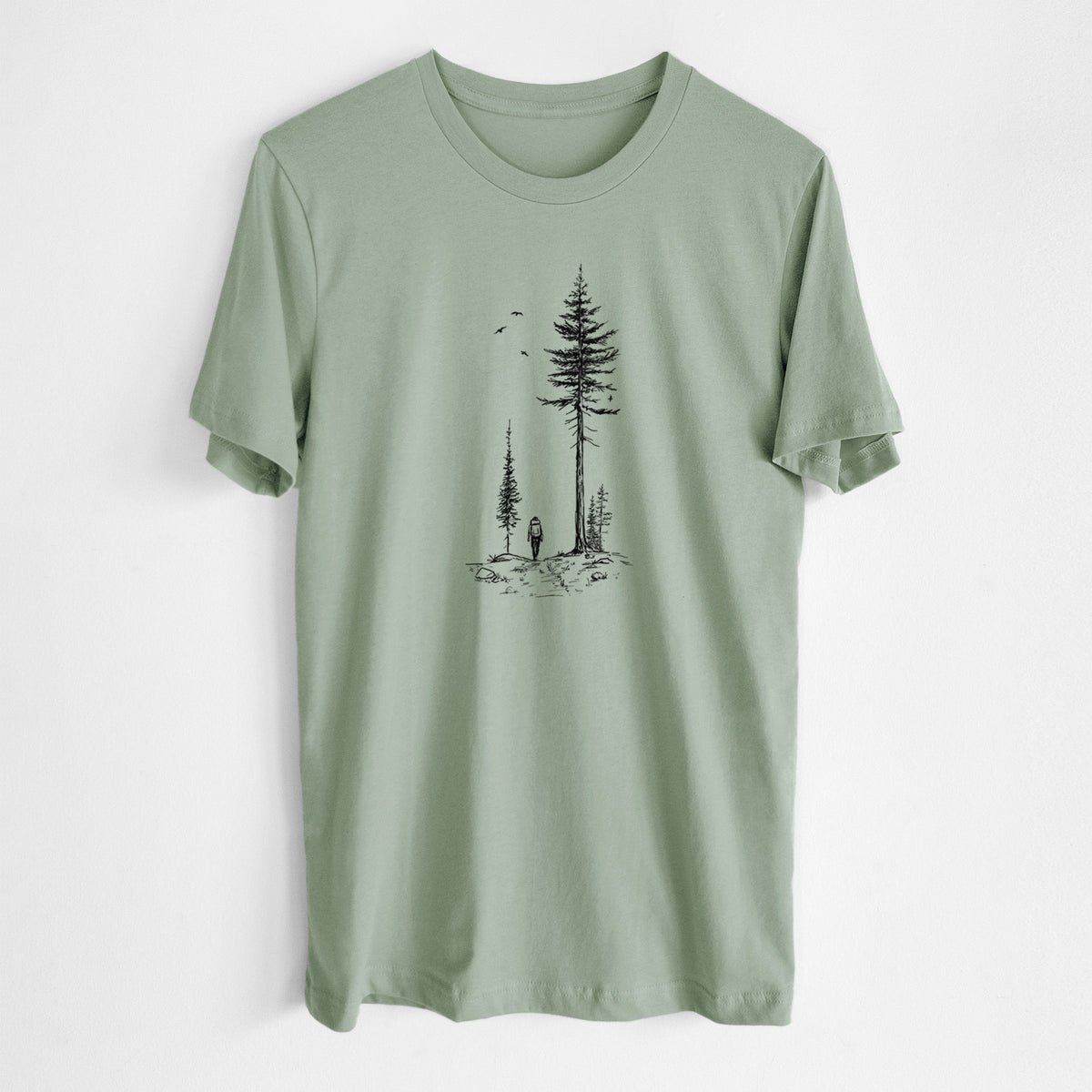Sage green lightweight cotton t-shirt laid flat, printed with a black ink illustration of a lone hiker walking between tall pine trees with birds overhead.