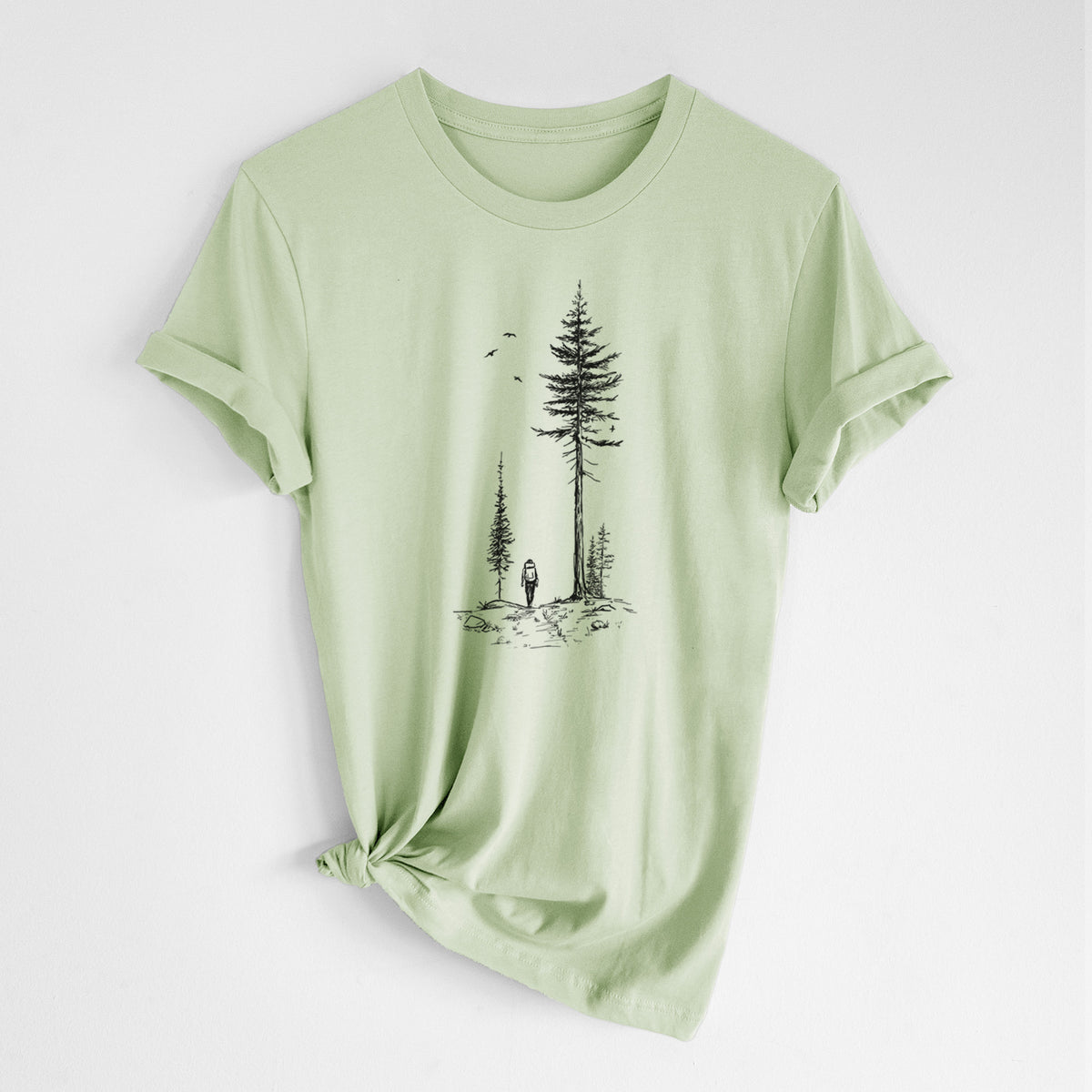 Spring green lightweight cotton t-shirt laid flat with the sleeves rolled, printed with a black ink illustration of a lone hiker walking between tall pine trees with birds overhead.