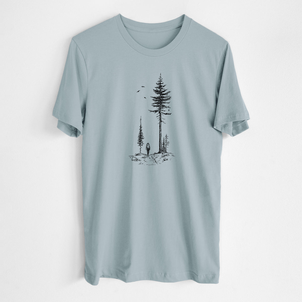 Vintage denim lightweight cotton t-shirt laid flat, printed with a black ink illustration of a lone hiker walking between tall pine trees with birds overhead.