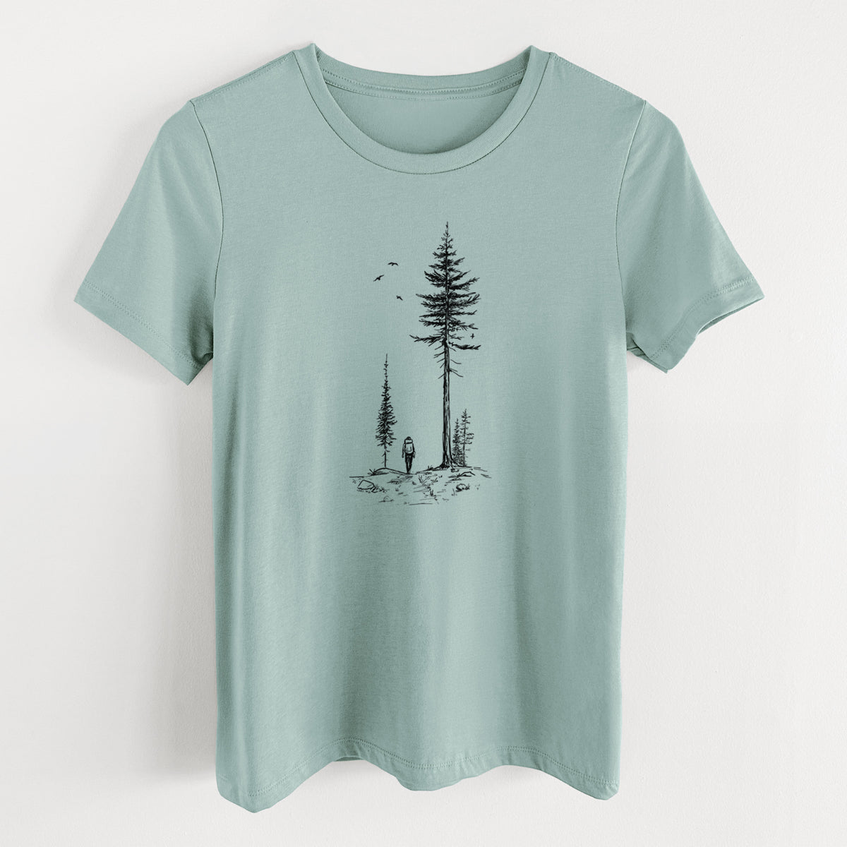 Dusty blue women's relaxed fit cotton t-shirt laid flat, printed with a black ink illustration of a lone hiker walking between tall pine trees with birds overhead.