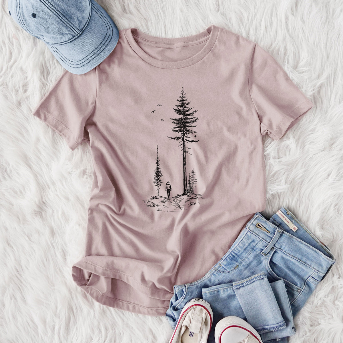 Women's relaxed fit cotton t-shirt printed with a black ink illustration of a lone hiker walking between tall pine trees with birds overhead, styled flat on a white faux fur rug with jeans.