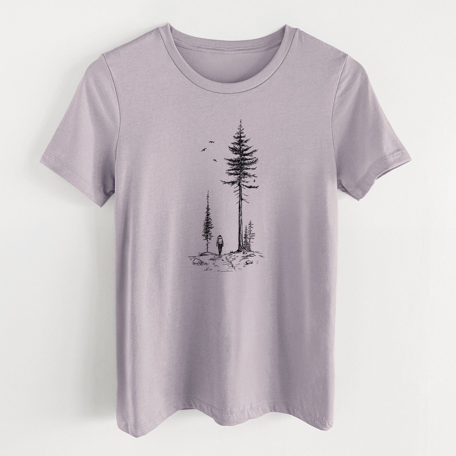 Light violet women's relaxed fit cotton t-shirt laid flat, printed with a black ink illustration of a lone hiker walking between tall pine trees with birds overhead.