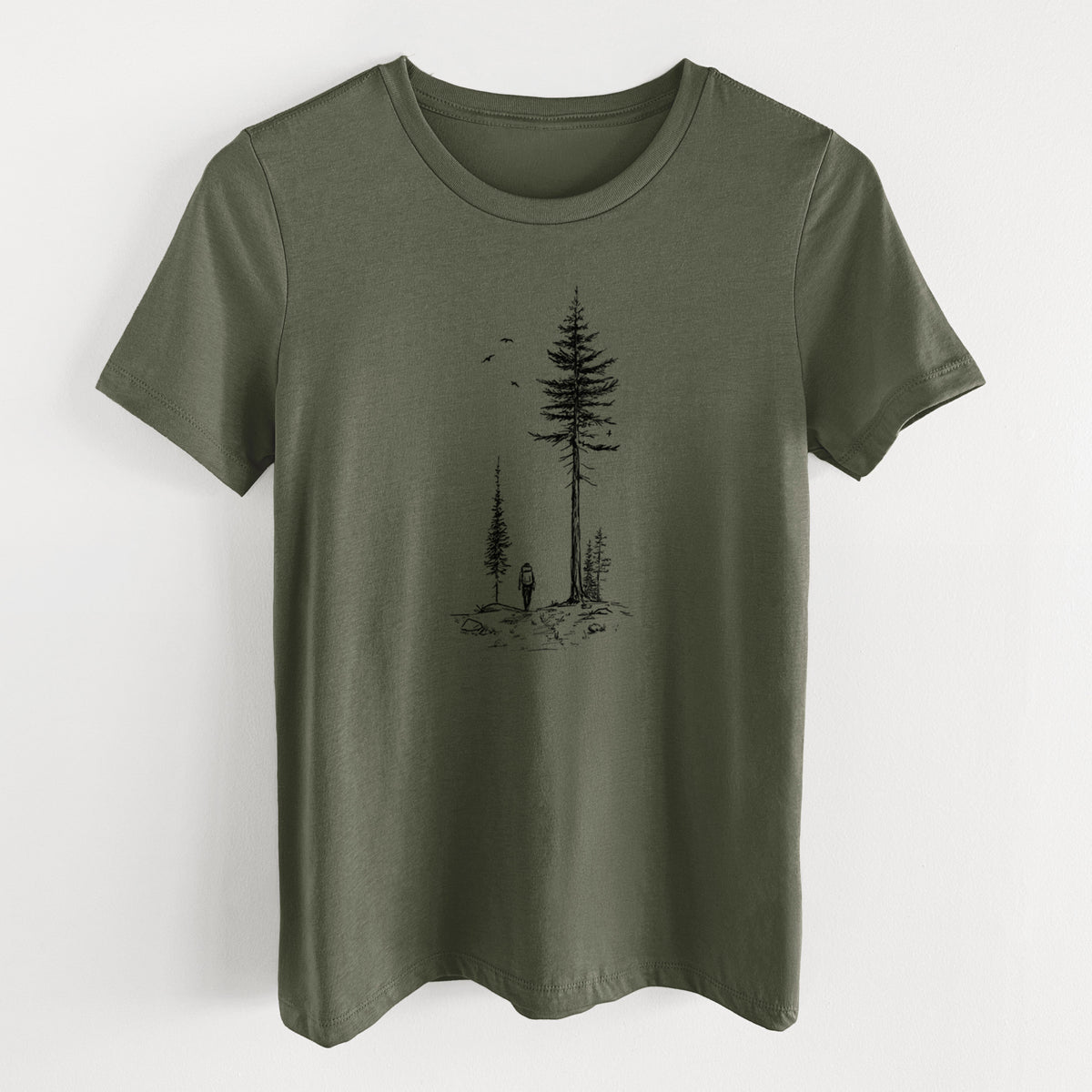 Military green women's relaxed fit cotton t-shirt laid flat, printed with a black ink illustration of a lone hiker walking between tall pine trees with birds overhead.