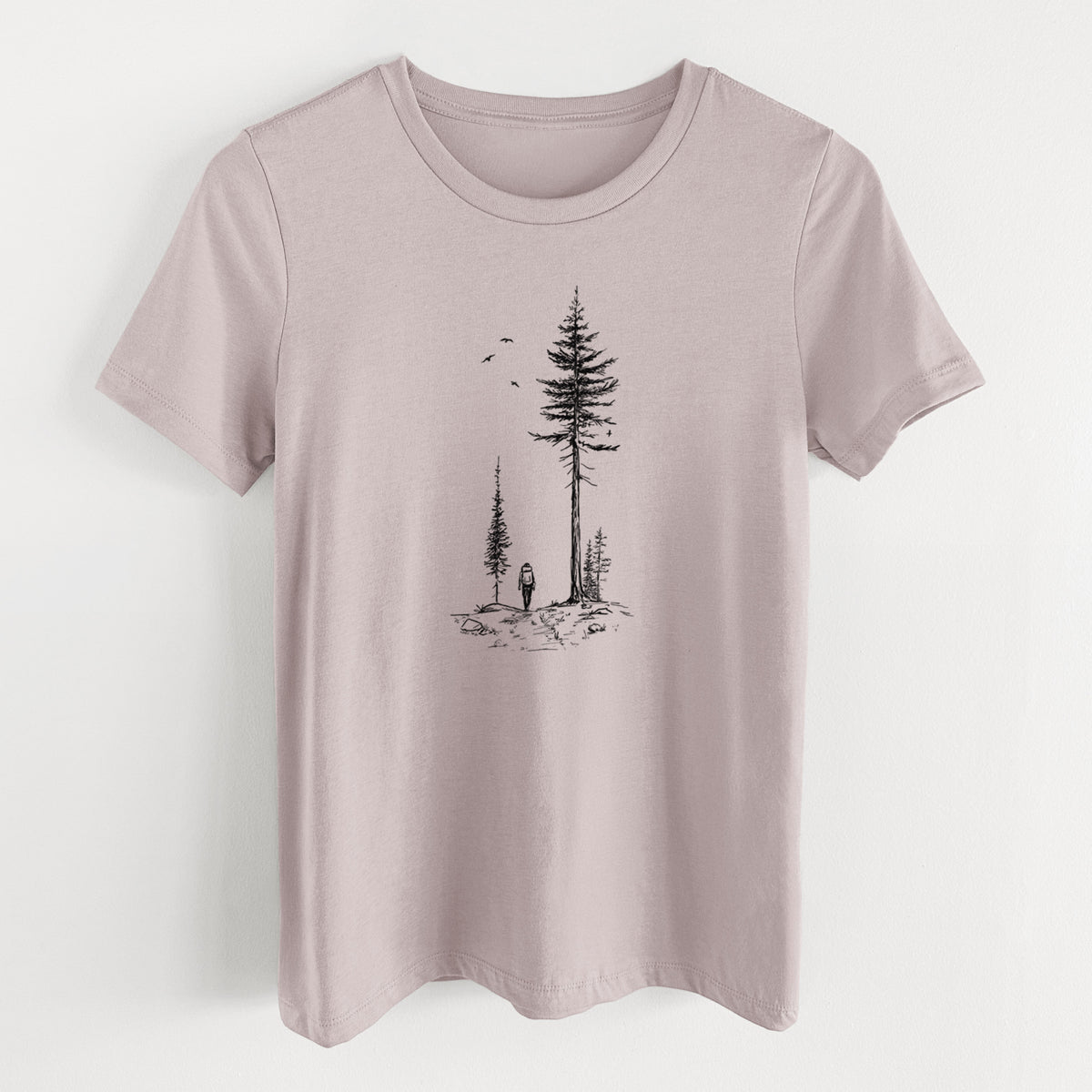 Pink gravel women's relaxed fit cotton t-shirt laid flat, printed with a black ink illustration of a lone hiker walking between tall pine trees with birds overhead.