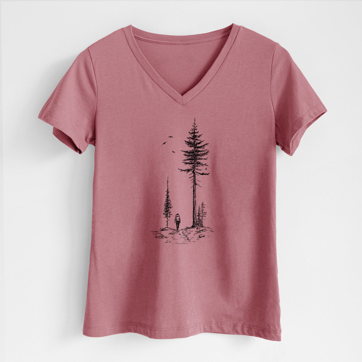 Mauve pink women's relaxed fit cotton V-neck t-shirt laid flat, printed with a black ink illustration of a lone hiker walking between tall pine trees with birds overhead.