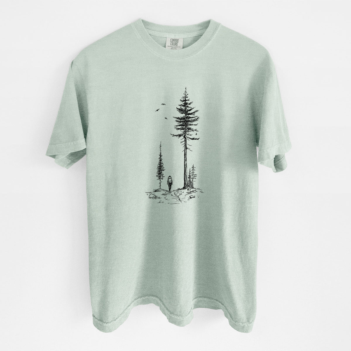 Bay green Comfort Colors t-shirt laid flat, printed with a black ink illustration of a lone hiker walking between tall pine trees with birds overhead.