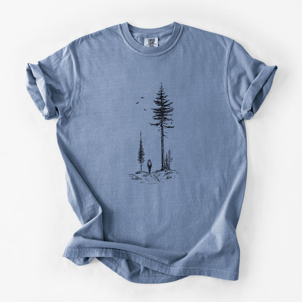 Blue jean Comfort Colors t-shirt laid flat, printed with a black ink illustration of a lone hiker walking between tall pine trees with birds overhead.