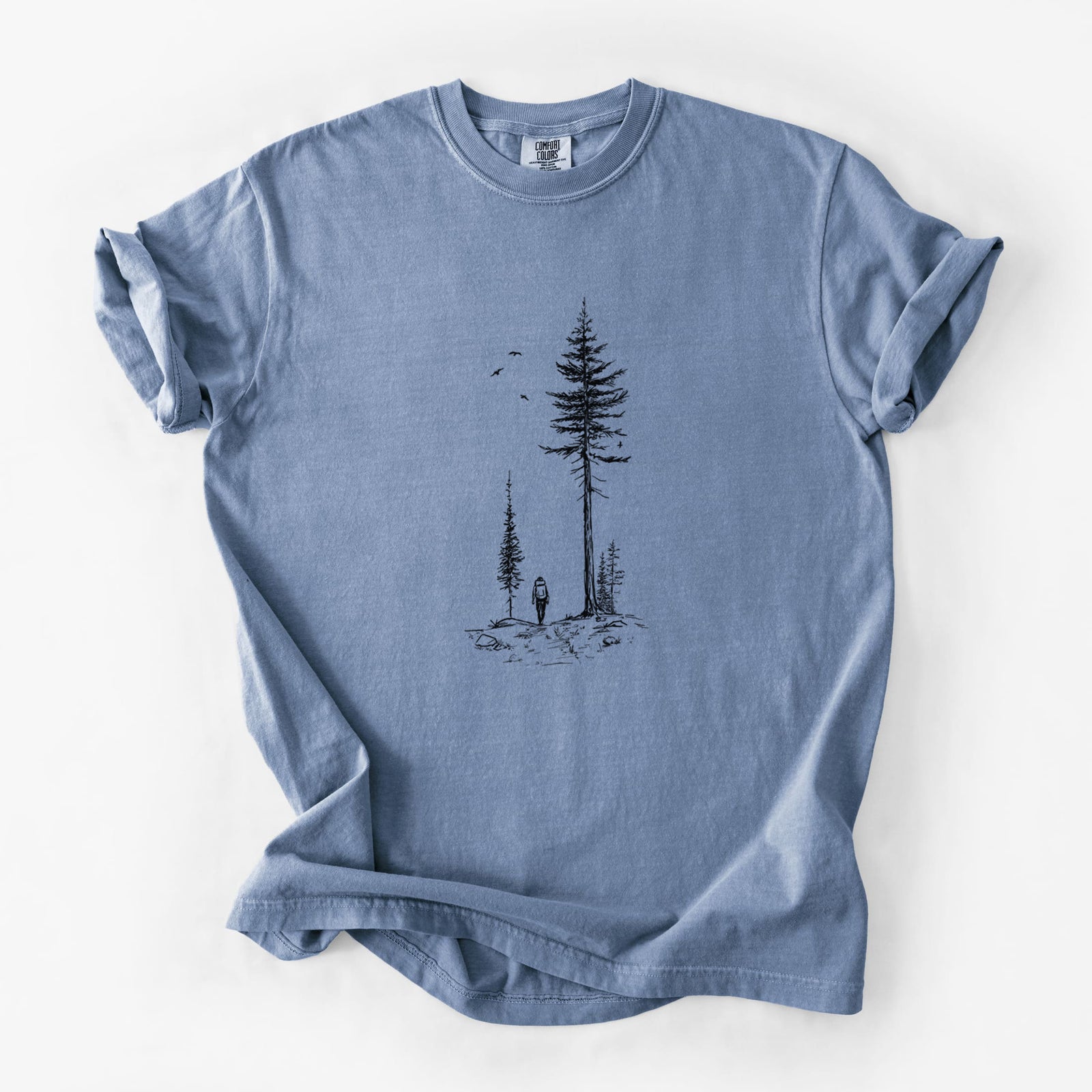 Blue jean Comfort Colors t-shirt laid flat, printed with a black ink illustration of a lone hiker walking between tall pine trees with birds overhead.