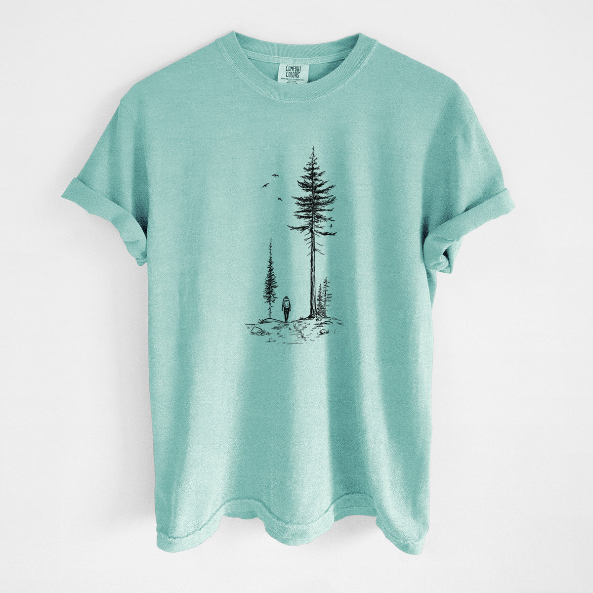 Chalky mint Comfort Colors t-shirt laid flat, printed with a black ink illustration of a lone hiker walking between tall pine trees with birds overhead.