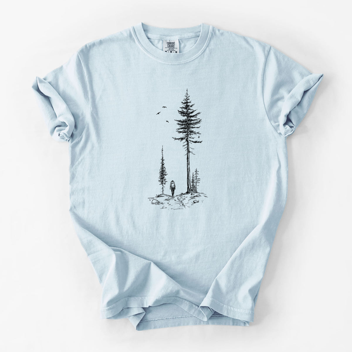 Chambray blue Comfort Colors t-shirt laid flat, printed with a black ink illustration of a lone hiker walking between tall pine trees with birds overhead.
