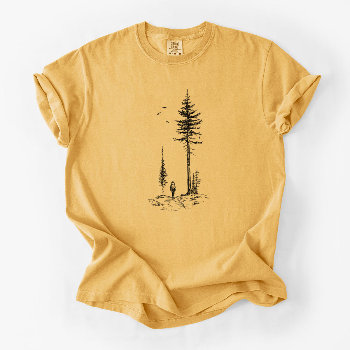 Golden wheat yellow Comfort Colors t-shirt laid flat, printed with a black ink illustration of a lone hiker walking between tall pine trees with birds overhead.