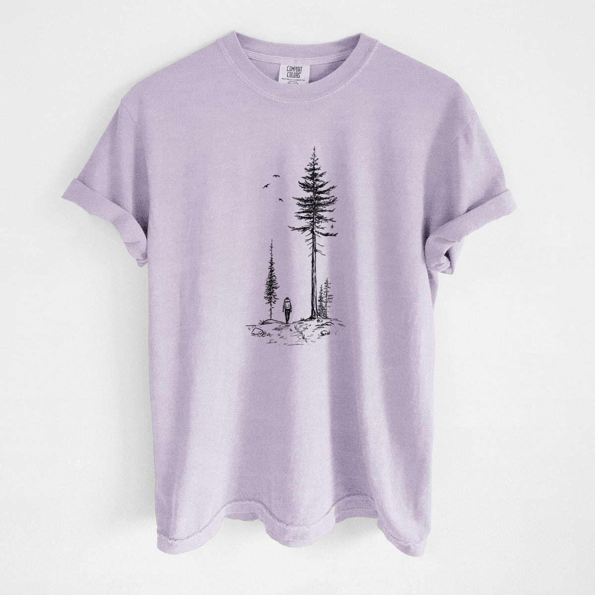 Orchid purple Comfort Colors t-shirt laid flat, printed with a black ink illustration of a lone hiker walking between tall pine trees with birds overhead.