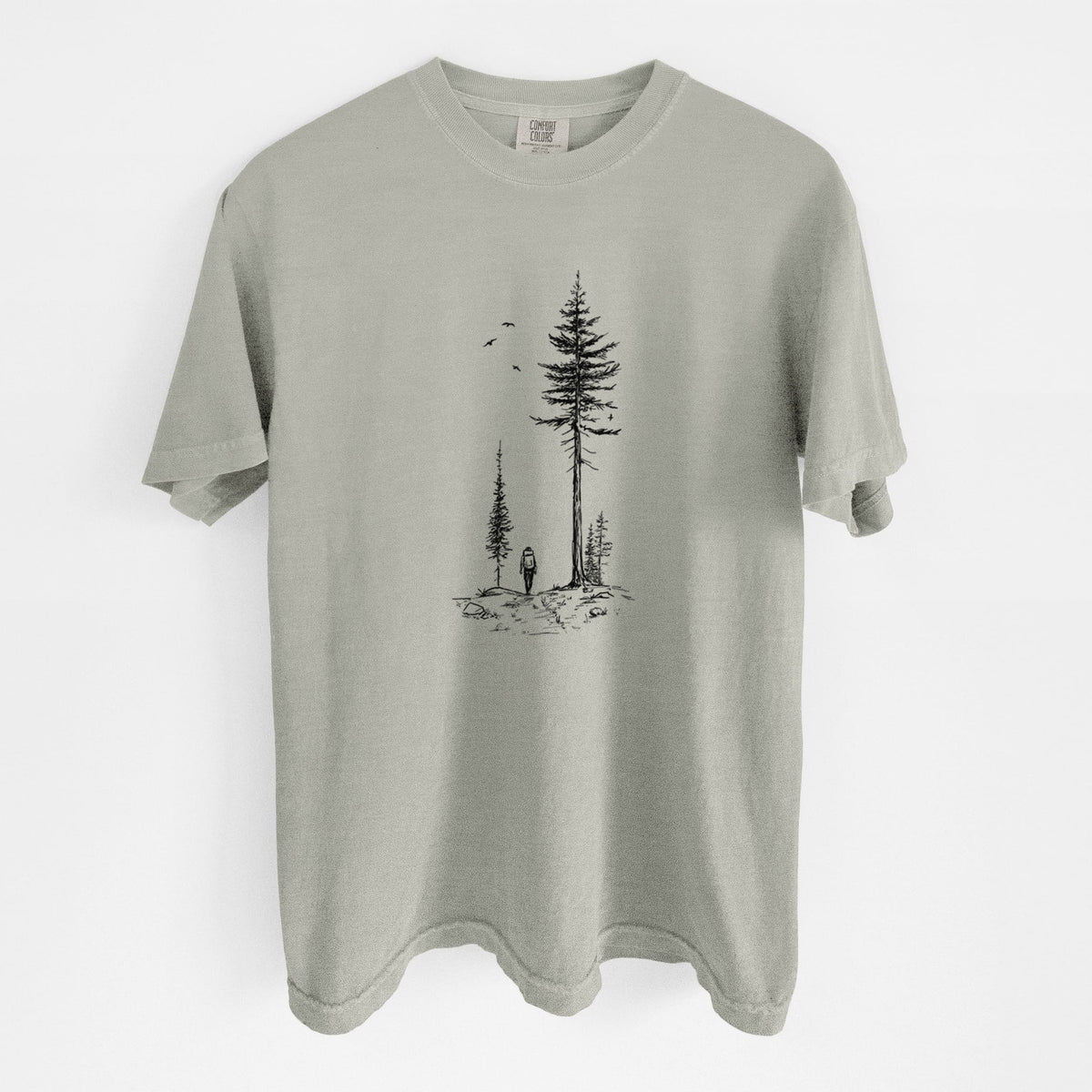 Sandstone beige Comfort Colors t-shirt laid flat, printed with a black ink illustration of a lone hiker walking between tall pine trees with birds overhead.