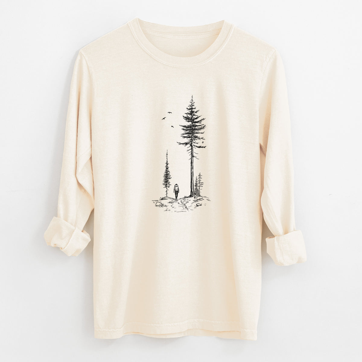 Ivory Comfort Colors long sleeve shirt laid flat with the cuffs rolled, printed with a black ink illustration of a lone hiker walking between tall pine trees with birds overhead.
