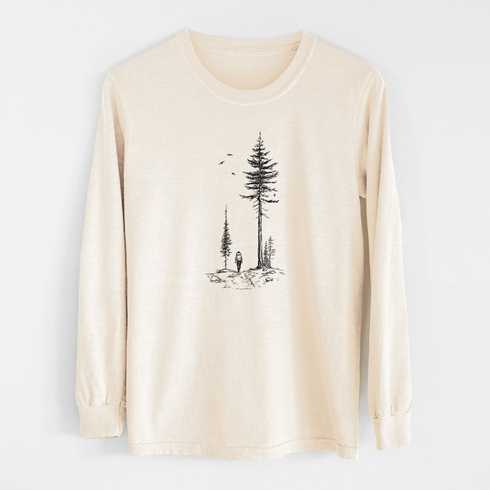 Ivory Comfort Colors long sleeve shirt laid flat, printed with a black ink illustration of a lone hiker walking between tall pine trees with birds overhead.