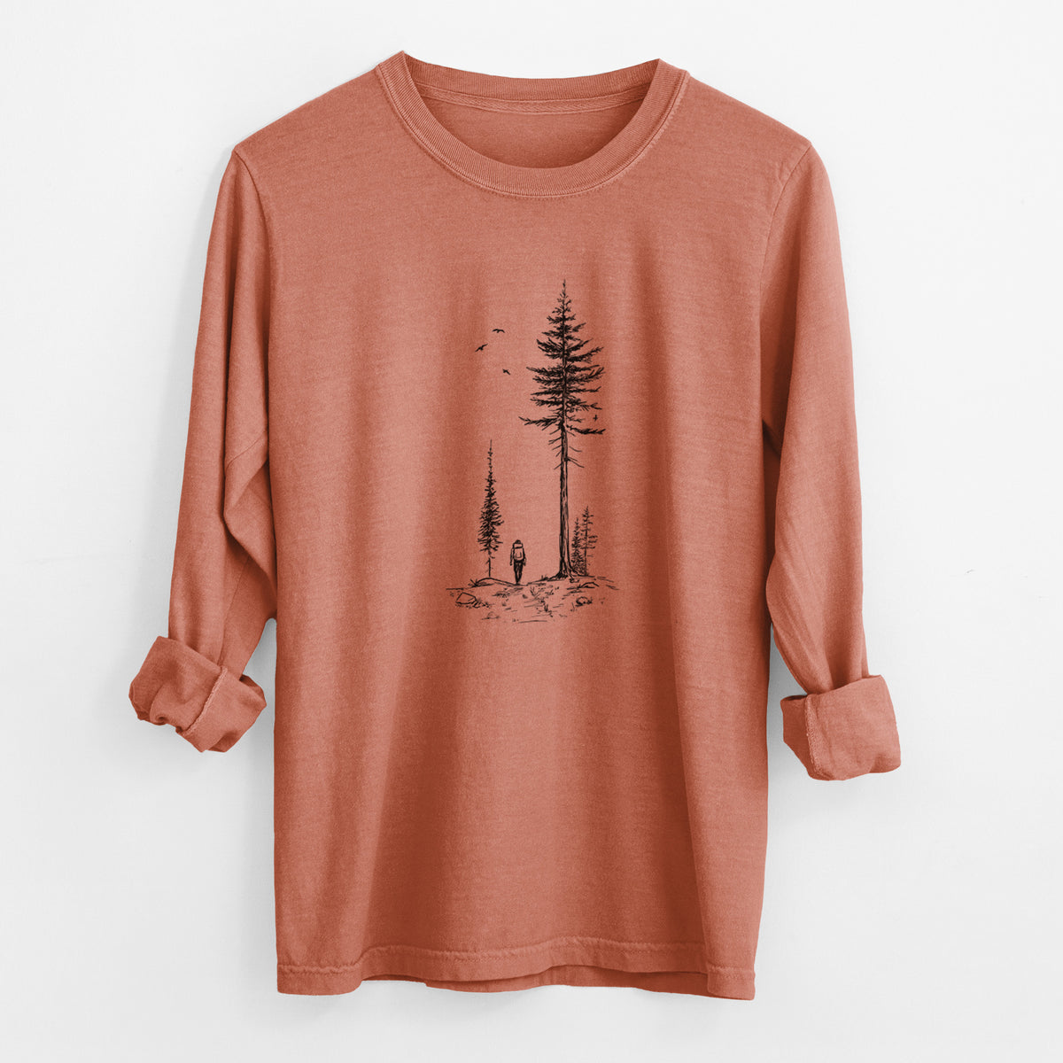 Yam burnt orange Comfort Colors long sleeve shirt laid flat with the cuffs rolled, printed with a black ink illustration of a lone hiker walking between tall pine trees with birds overhead.