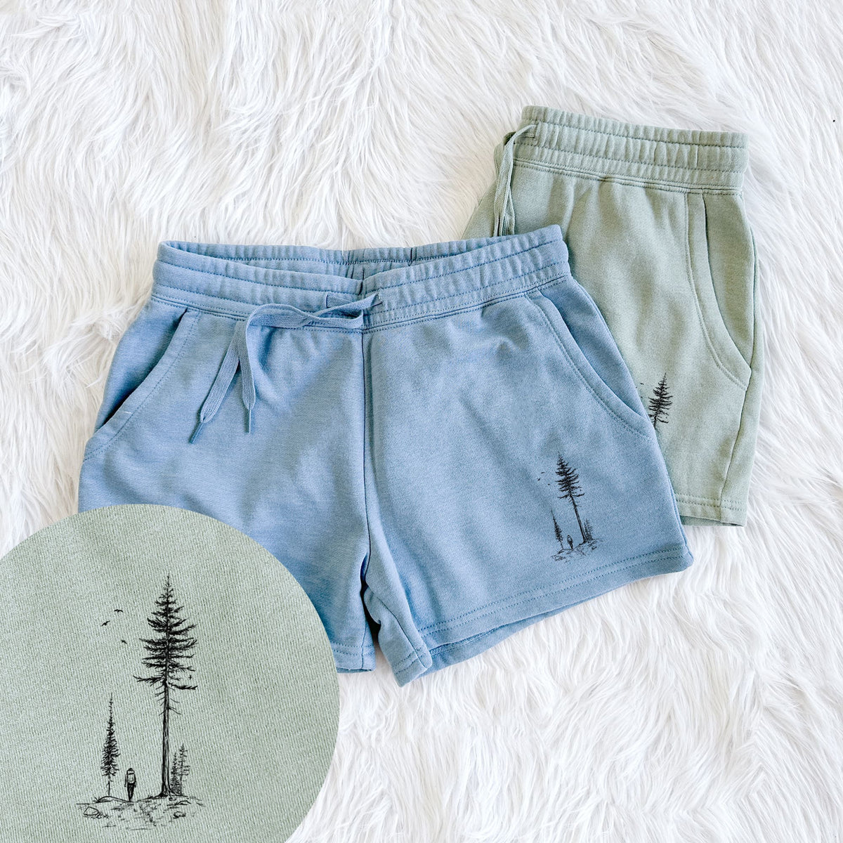 Two pairs of Cali Wave shorts in different colors printed with a black ink illustration of a lone hiker walking between tall pine trees with birds overhead, with a close-up of the print.