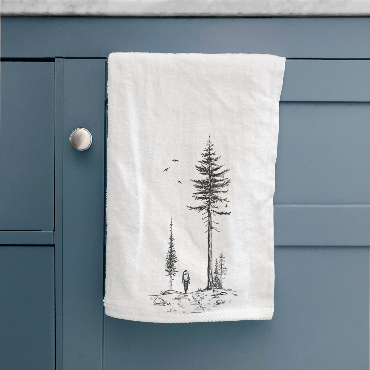 White hand towel printed with a black ink illustration of a lone hiker walking between tall pine trees with birds overhead, hanging on a cabinet door.
