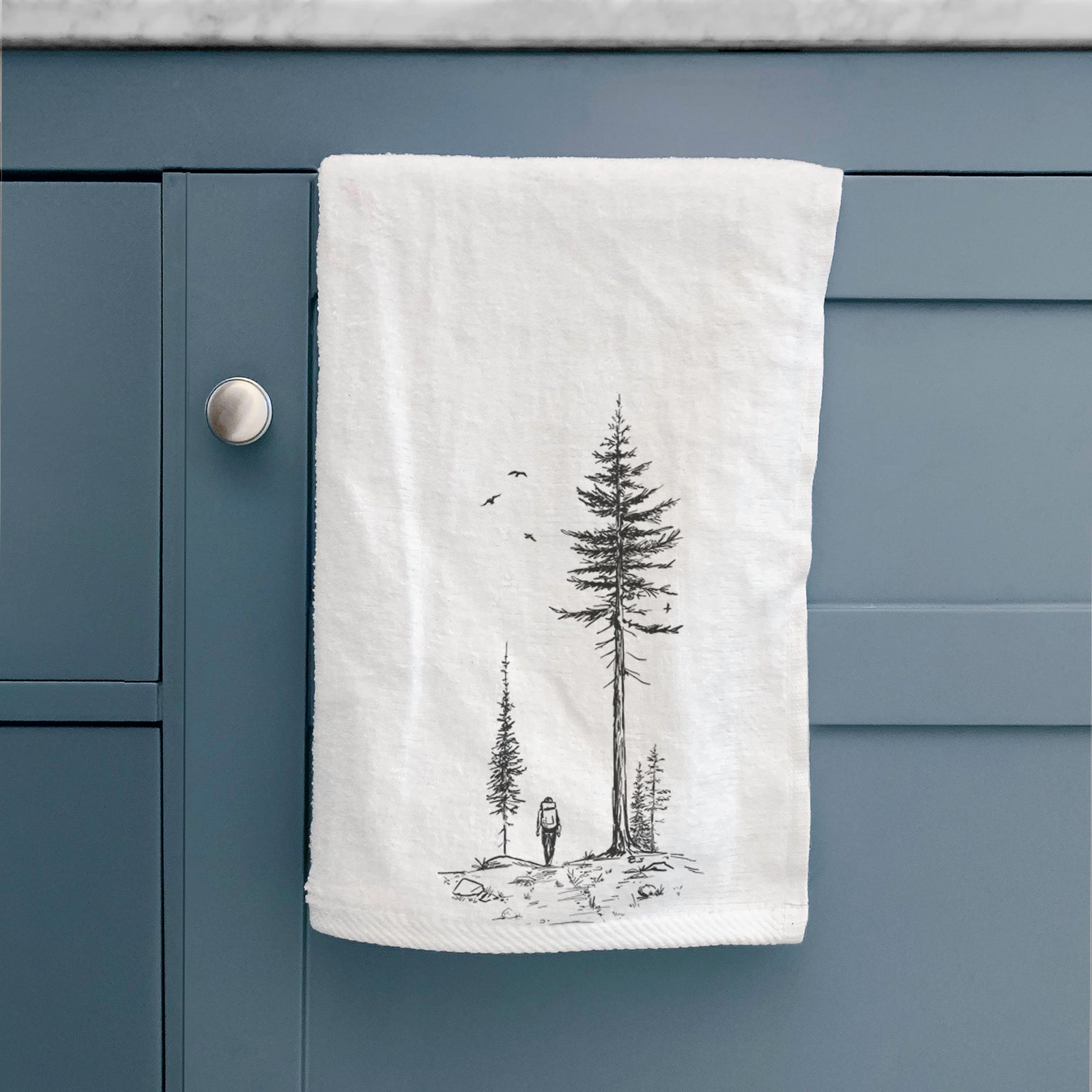 White hand towel printed with a black ink illustration of a lone hiker walking between tall pine trees with birds overhead.